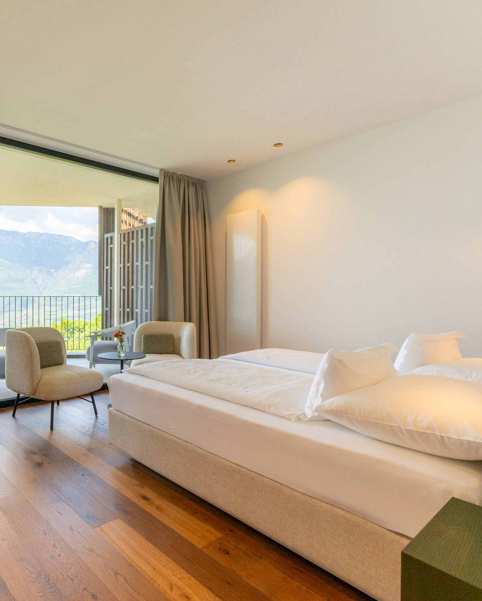 Bright hotel room with a double bed, two chairs, and a balcony overlooking mountains and greenery. - Hotel Tenz