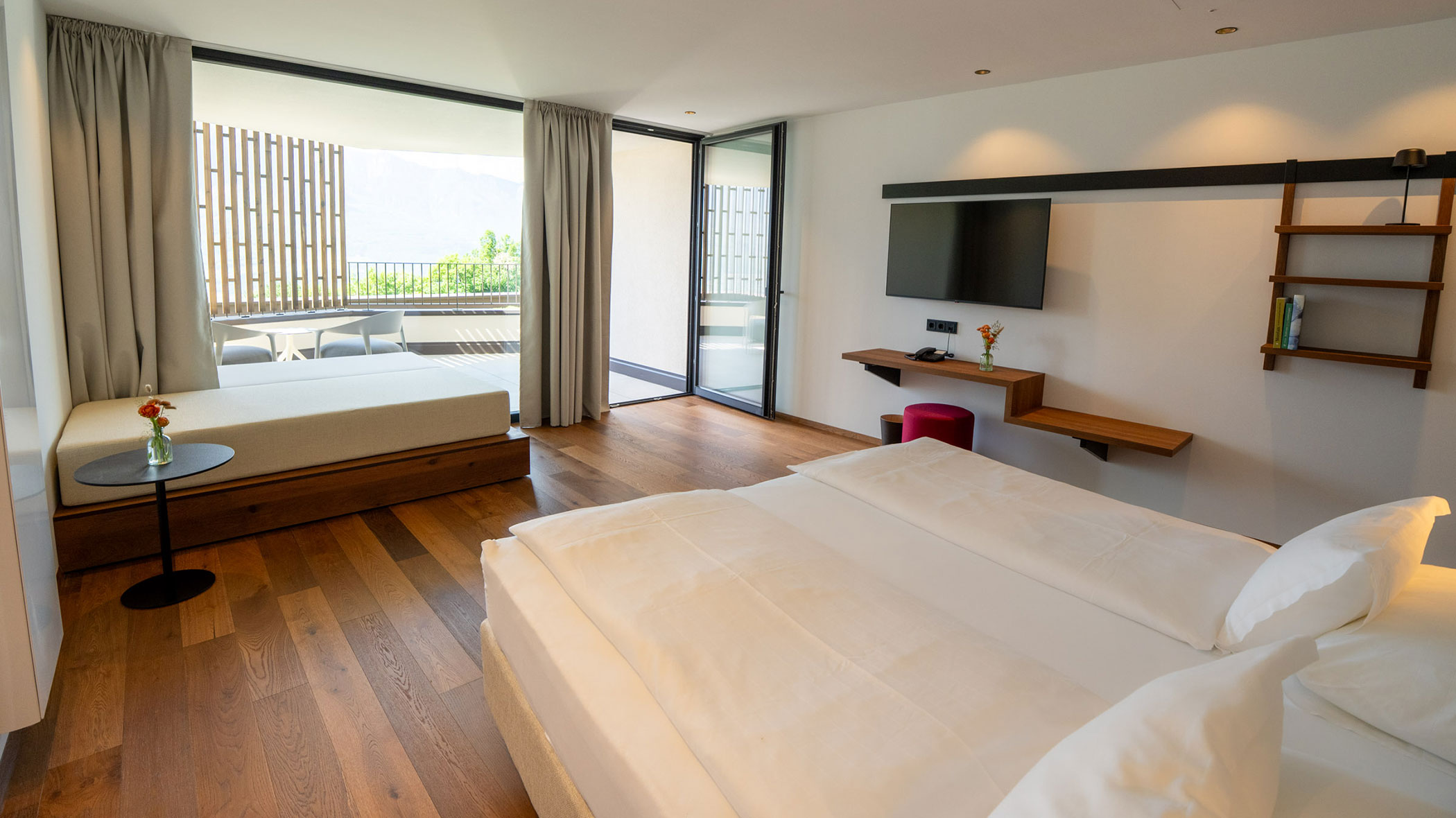 Modernes Hotelzimmer mit Doppelbett, Schlafsofa, TV, Balkon, Holzboden und großen Fenstern, die viel Tageslicht hereinlassen. - Hotel Tenz